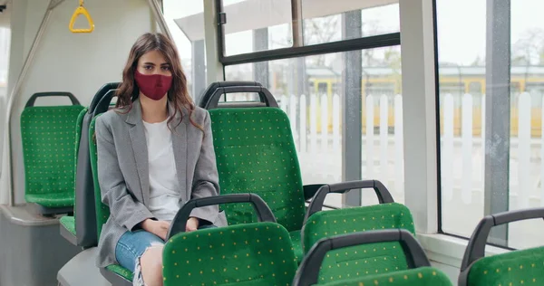 Retrato de niña en máscara de algodón sentado en el autobús vacío y mirando a la cámara. Mujer morena milenaria en ropa casual con máscara protectora reutilizable. Concepto de virus y pandemia . Imágenes de stock libres de derechos