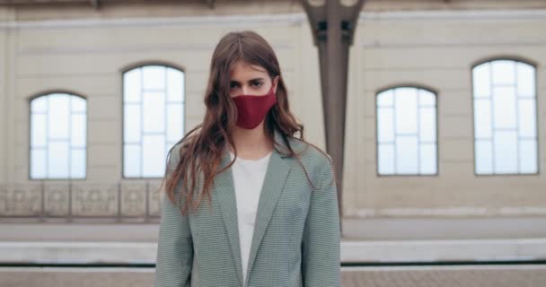 Ritratto di ragazza bruna millenaria in abiti casual in piedi sulla piattaforma vuota della stazione ferroviaria. Vista Crop di giovane donna di tendenza in maschera facciale fatta a mano. Concetto di pandemia virale . — Video Stock