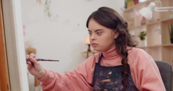 Close-up uitzicht op schattige talanted teenager schilderij schilderij terwijl zitten in de voorkant van molbert. mooi meisje met genetische stoornis dragen in kunstenaar aprone en het doen van kunst in haar kamer. — Stockvideo