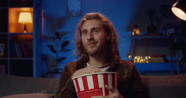 Un joven guapo comiendo palomitas y viendo películas en casa. Mullennial chico ingenio barba y pelo largo sentado en el sofá delante de la televisión y sonriendo mientras pasar el tiempo en la noche . — Vídeos de Stock