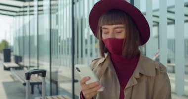 Demiryolu istasyonunda kürsüde otururken akıllı telefon kullanan pamuk protektif maskeli bir kadını yakından çek. Bavullu şık elbiseli bayan yolcu telefon ekranına bakıyor..