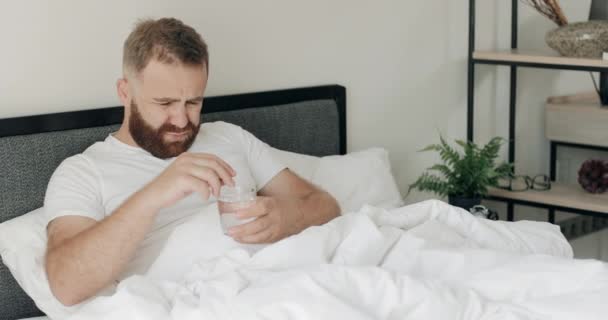 젊은 턱수염 남자가 독극물 치료제를 물 잔에 던지고 있어. 30 대의 친절 한 남자가 침대에 누워 밤새 술을 마시고 숙취에 시달리고 몸 이 안 좋은 것을 보고 있습니다. — 비디오