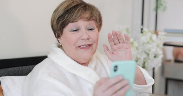 Vista de cerca de la anciana sonriendo mientras recibe una videollamada emocional con su hijo o hija. Hermosa anciana vistiendo roba casa hablando y haciendo gestos mientras está sentada en la cama . — Vídeos de Stock