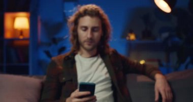 Uzun saçlı hippi adamın portresi akıllı telefon ekranına bakıyor ve kafasını kaldırıyor. Mobil telefon tutarken kameraya gülümseyen ve evdeki kanepede oturan yakışıklı genç adam. Yakınlaştır.