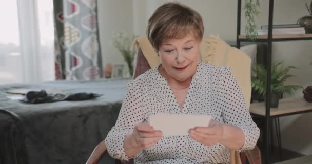 Mulher feliz olhando para fotos enquanto se senta na cadeira em casa.Idosos bela senhora nos anos 60 ter nostalgia ao passar por fotos. Conceito de memórias e emoções . — Vídeo de Stock