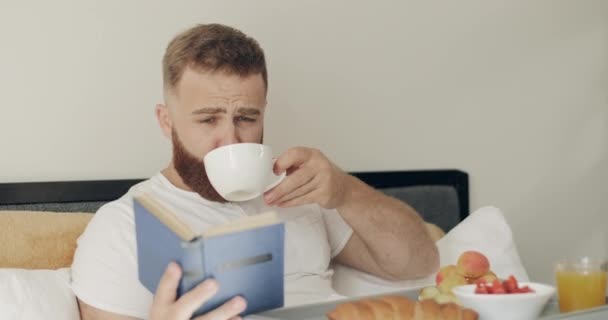 Hombre de 30 años bebiendo té y leyendo detective mientras está sentado con la bandeja llena de comida en sus piernas. Intelectual chico disfrutando de libro mientras desayuna en la cama. Concepto de ocio . — Vídeo de stock