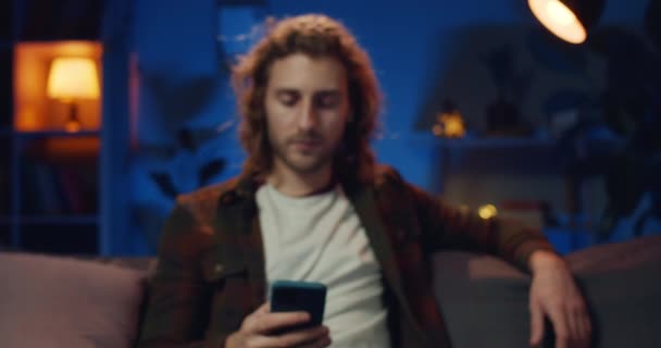 스마트폰 화면을 보고 머리를 들고 있는 긴 머리의 히프 스터 남자의 초상화입니다. 휴대 전화를 들고 소파에 앉아 카메라 앞에서 미소짓고 있는 젊은 청년. 줌 인. — 비디오