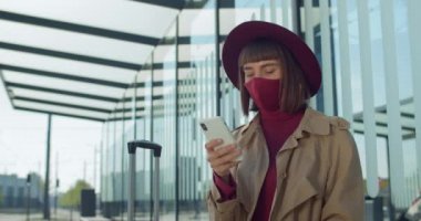 Kız bankta otururken ve akıllı telefon kullanırken yakın çekim. Pamuk maskeli kadın ayağa kalkıp tren istasyonunun yanında bavul taşıyor. Koronavirüs ve seyahat kavramı.