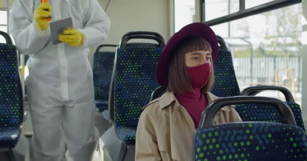 Millennial meisje zit in de trein terwijl de temperatuur te controleren. Medische medewerker met tablet en thermometr draagt beschermende pak screening vrouwelijke passagier. Concept van Covid-19. — Stockvideo