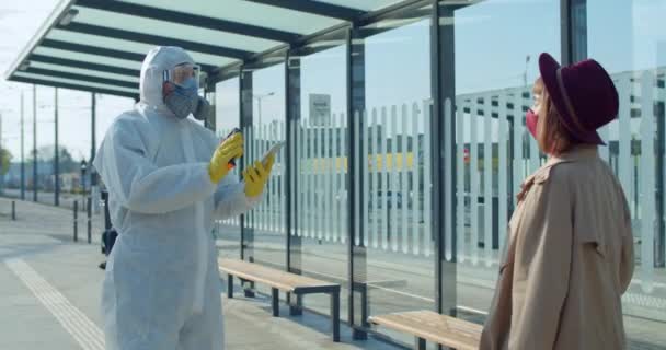 Viajera milenaria que tiene control de temperatura cerca del aeropuerto. Trabajador médico con traje protector usando tableta y termómetro mientras inspecciona al pasajero. Concepto de epidemia de Covid . — Vídeo de stock