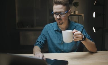 Elinde bardak olan adam dizüstü bilgisayara bakıyor.