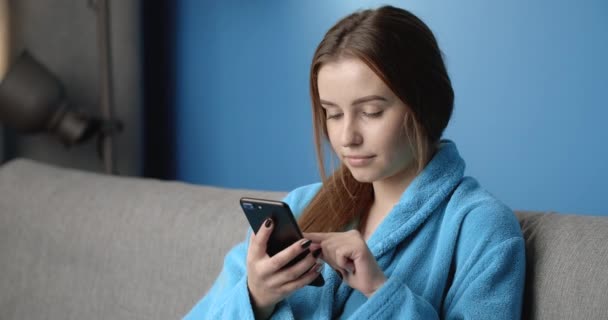 Mooie dame in blauwe badjas ontspannen op de bank met telefoon — Stockvideo