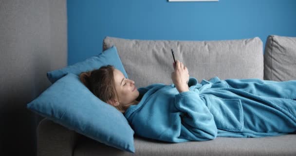 Gelukkig meisje in azuurblauwe badjas liggend op bank met mobiele telefoon — Stockvideo