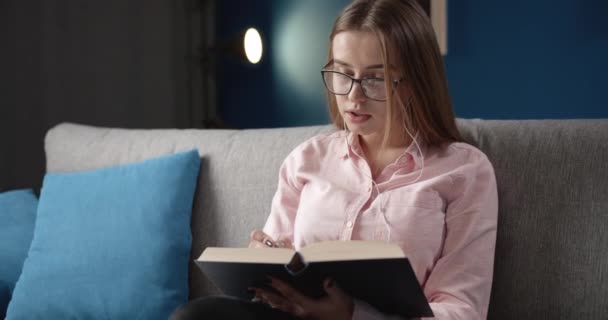 Menina bonita em fones de ouvido leitura livro em voz alta no sofá — Vídeo de Stock