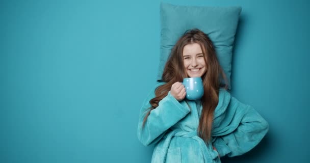 Сонная девушка в синем халате пьет кофе, чтобы проснуться — стоковое видео