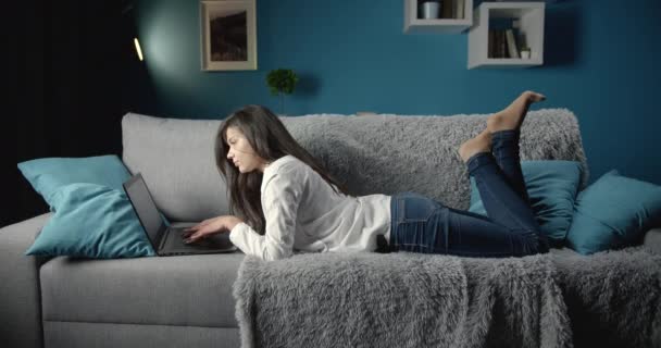 Женщина в повседневной одежде лежит на диване с ноутбуком — стоковое видео