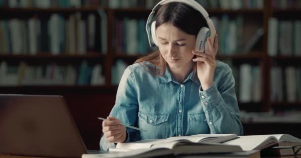 Студентка в наушниках, читающая в библиотеке — стоковое видео
