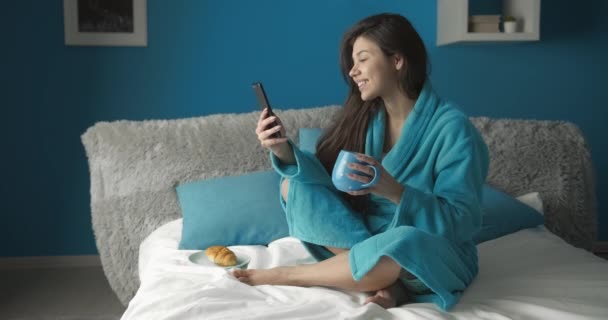 女性はベッドでコーヒーとスクロールスマートフォンを飲む — ストック動画