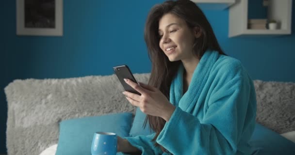 Frau trinkt Kaffee und blättert Smartphone im Bett — Stockvideo