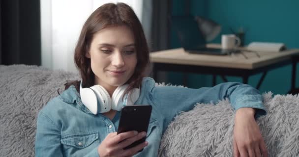 Alegre dama sentada en casa con auriculares y smartphone — Vídeo de stock