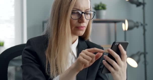 Γυναίκα με αυτοπεποίθηση που κάθεται στο γραφείο και χρησιμοποιεί smartphone — Αρχείο Βίντεο
