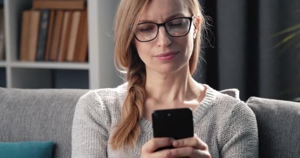 Glad kvinna sitter på soffan och sms: ar på smartphone — Stockvideo