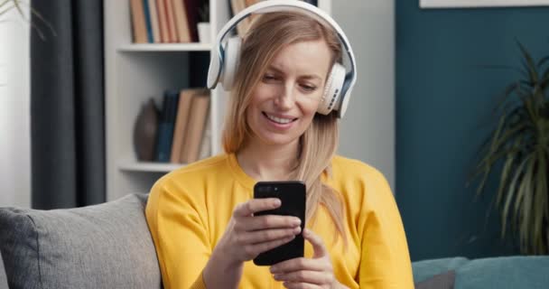 Happy female sitting with headphones and smartphone on couch — Stock Video