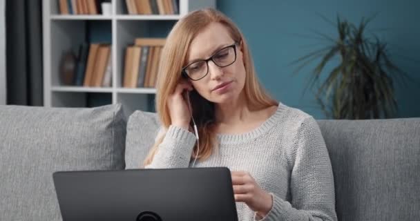Ώριμη κυρία που χρησιμοποιεί ακουστικά για την εργασία σε φορητό υπολογιστή στο σπίτι — Αρχείο Βίντεο