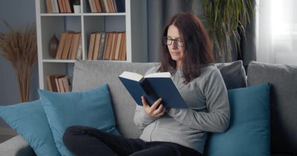 Χαρούμενη κυρία με γυαλιά ηλίου που διαβάζει βιβλίο στον καναπέ — Αρχείο Βίντεο