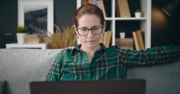 Χαλαρή γυναίκα κάθεται στον καναπέ και εργάζεται σε φορητό υπολογιστή — Αρχείο Βίντεο