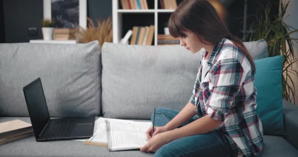 Tampilan samping gadis muda melakukan pekerjaan rumah dengan laptop dan buku — Stok Video