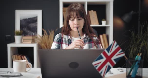 Девушка сидит за столом с ноутбуком и изучает английский язык — стоковое видео