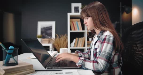 Вид сбоку девушки, сидящей за столом и сидящей на ноутбуке — стоковое видео