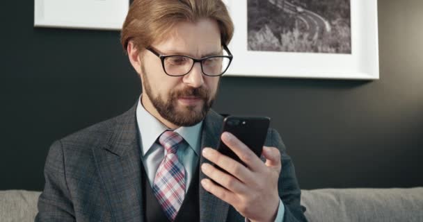 Portret van een man met baard die op de bank zit en een smartphone gebruikt — Stockvideo