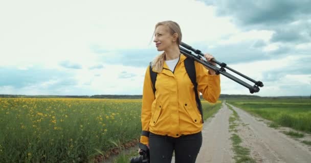 Женщина ходит по полю с фотоаппаратом и штативом — стоковое видео