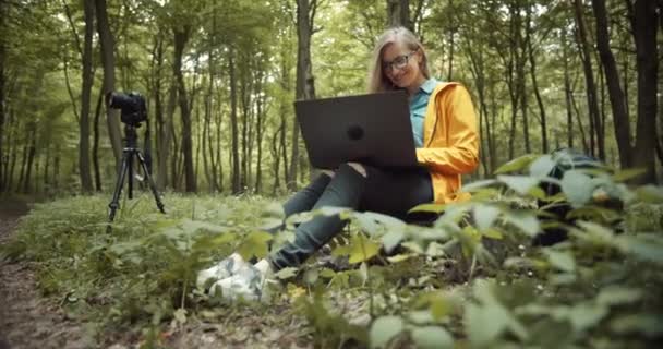 Feliz fotógrafo trabajando en el ordenador portátil mientras está sentado en el bosque — Vídeos de Stock
