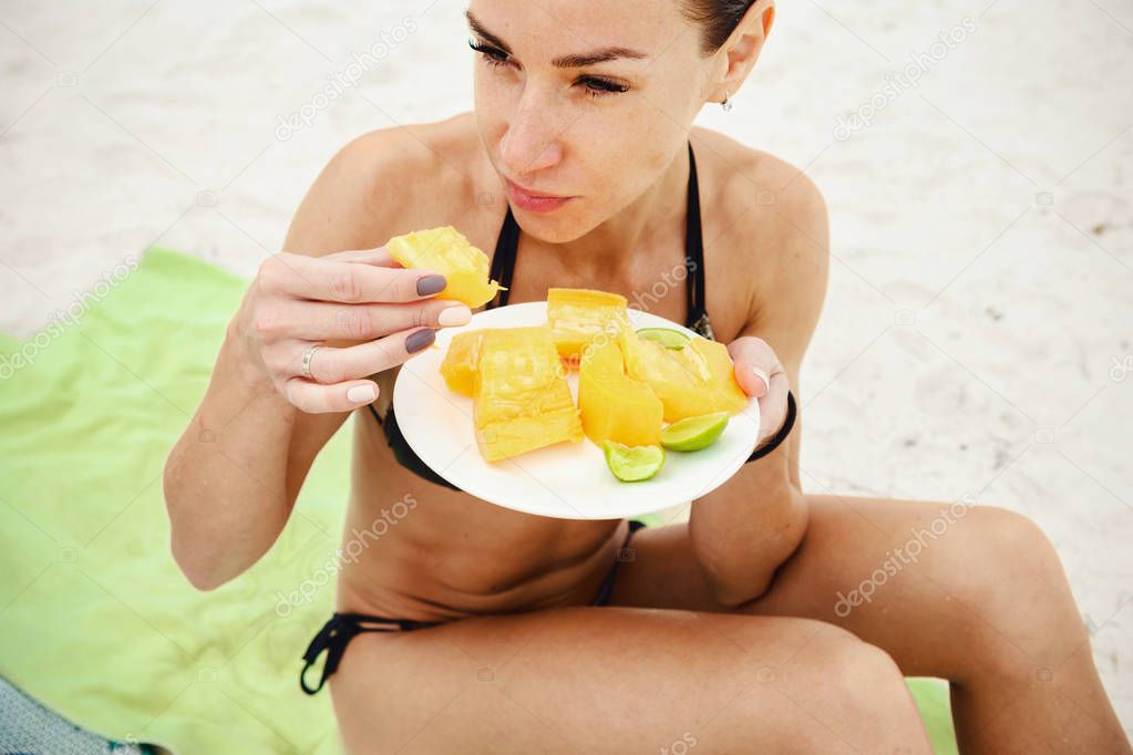 Young woman sitting on beach eating papaya