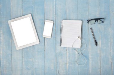 Çalışma alanı ile beyaz tablet, akıllı telefonlar, not defteri, kulaklık, kalem ve gözlük ahşap mavi masa, üstten görünüm