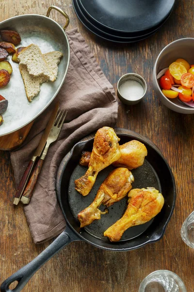 Dinner table concept. Fried chicken legs in frying pan on wooden table with potatoes and salad
