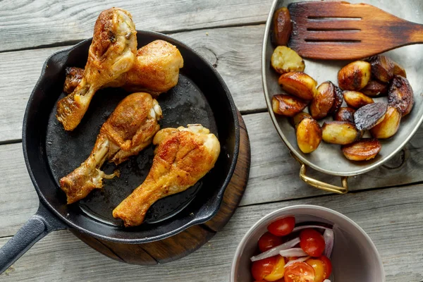 Fried chicken legs with fried potatoes in pan on wooden table with salad, top view