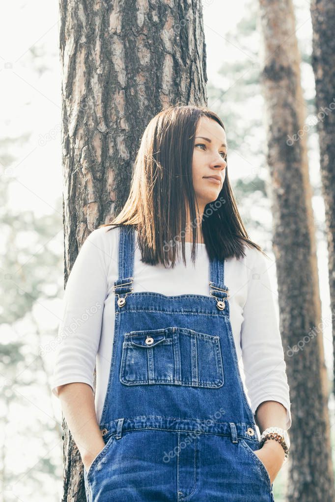 Beautiful young woman in jeans overalls standing in woodland at sunny day
