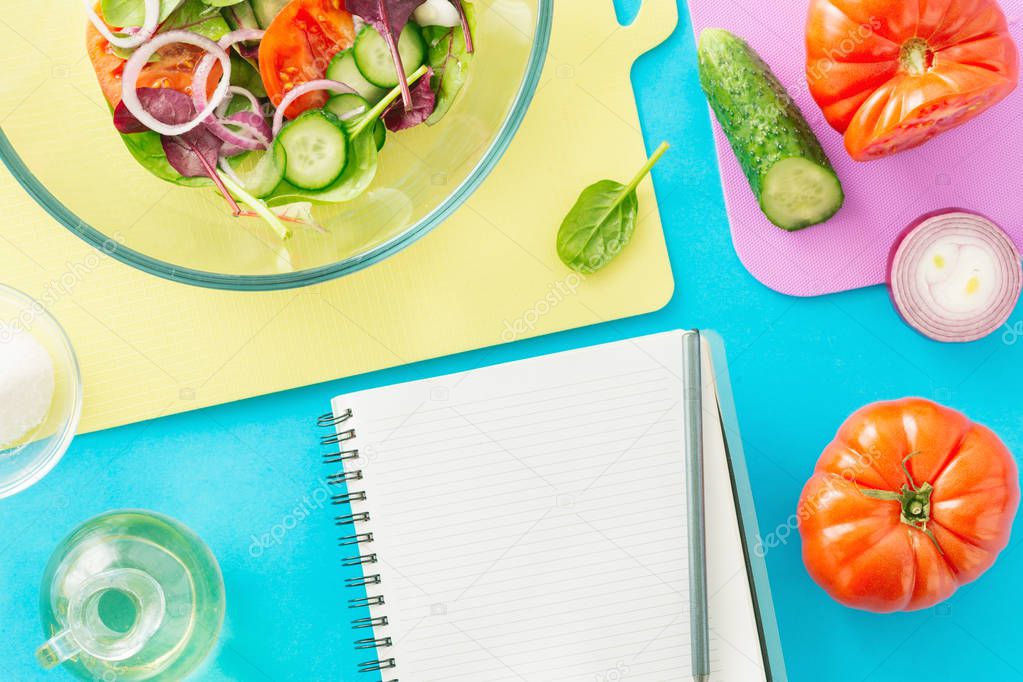 Notepad with fresh biodynamic ingredients for cooking summer salad on blue background, top view, Healthy food concept