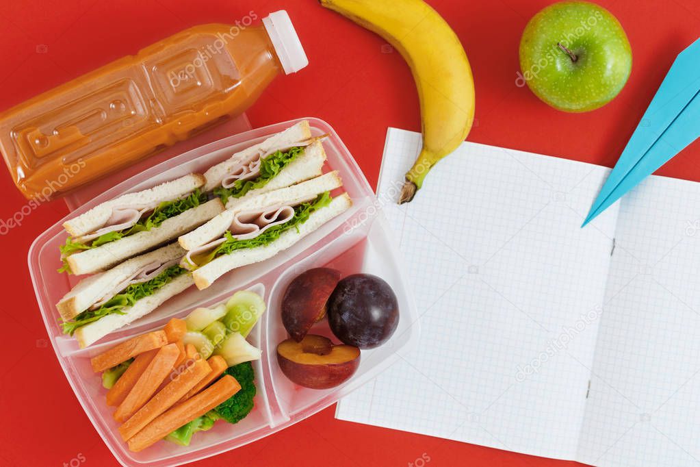Notebook with blank pages with healthy school lunch boxes with sandwiches, fresh vegetables and bottle of juice on red background
