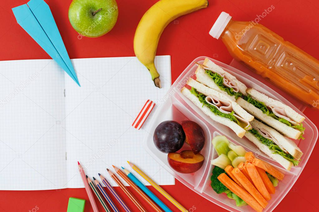 Notebook with blank pages with healthy school lunch boxes with sandwiches, fresh vegetables, bottle of juice and school supplies on red background