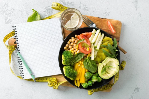 Vegan sağlıklı dengeli diyet konsepti. Boş defter ve mezura ile vejetaryen Buda kasesi. Beyaz arka planda bezelye, brokoli, biber, domates, ıspanak, roka ve avokado tabağı. Üst görünüm 