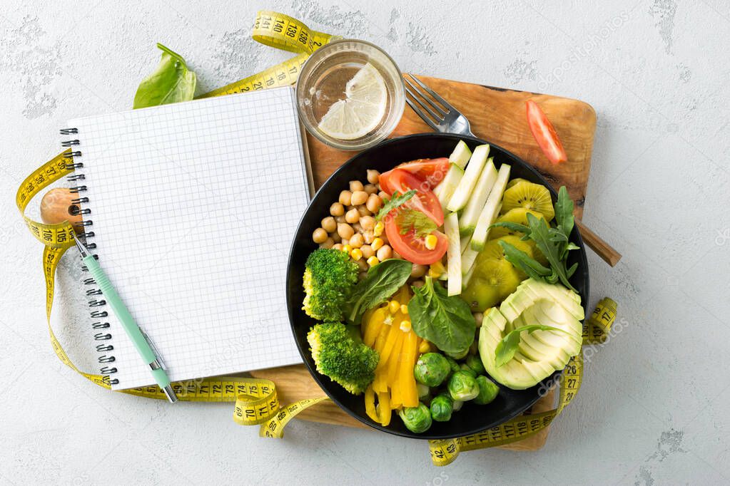 Vegan healthy balanced diet concept. Vegetarian buddha bowl with blank notebook and measuring tape. hickpeas, broccoli, pepper, tomato, spinach, arugula and avocado in plate on white background. Top view 