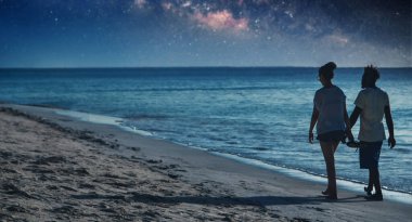 Birbirine aşık çiftler gün batımında okyanus kıyısında el ele yürüyorlar.