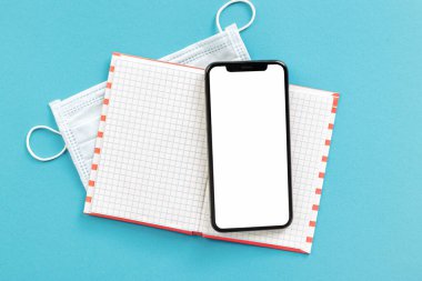 Mavi arkaplan arkaplanında akıllı telefon ve tıbbi yüz maskesi olan bir defter. Uzak çalışma kavramı.