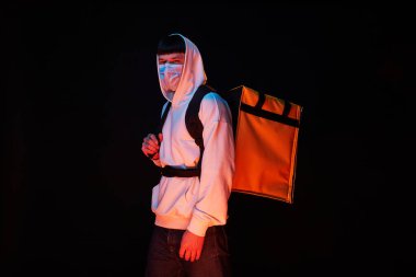 Siyah arka planda sarı sırt çantalı, beyaz maskeli genç teslimatçı.