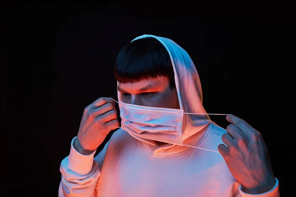 portrait young fashion guy in white hoodie in the hood robe in medicine mask in color lights on black background